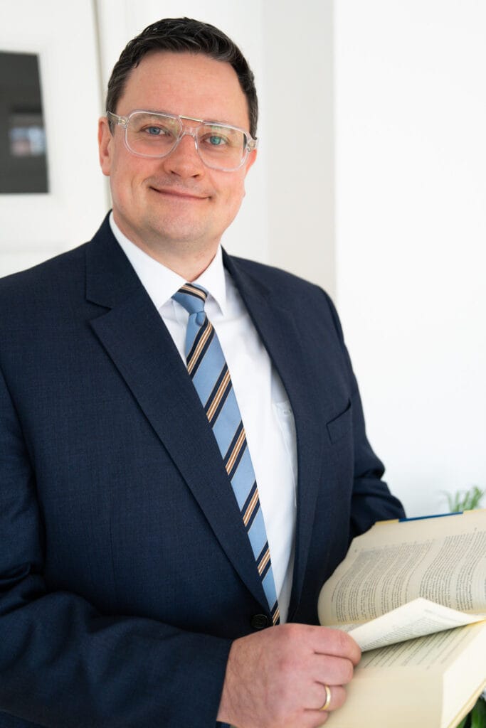 Ein lächelnder Mann mit Brille, Anzug und gestreifter Krawatte hält ein aufgeschlagenes Buch in seinen Händen. Er steht vor einem hellen Hintergrund.