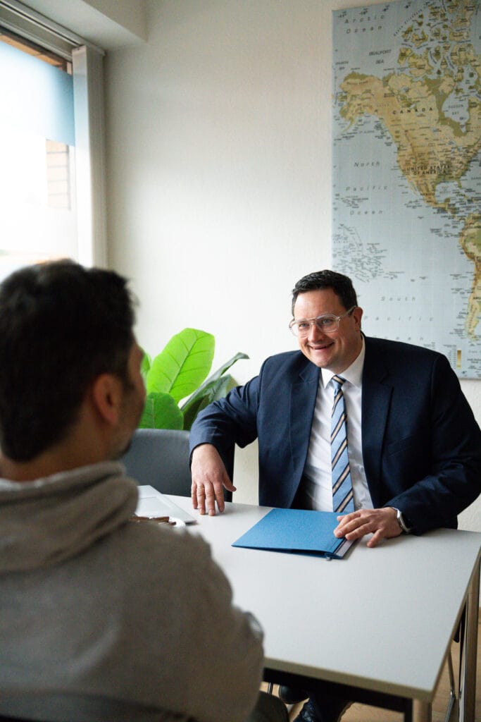 Ein Mann in einem dunkelblauen Anzug und einer gestreiften Krawatte sitzt an einem Tisch und lächelt. Er schaut einen anderen Mann an, der ihm gegenübersitzt und dessen Rücken zur Kamera zeigt. Im Hintergrund ist eine große Weltkarte an der Wand zu sehen.
