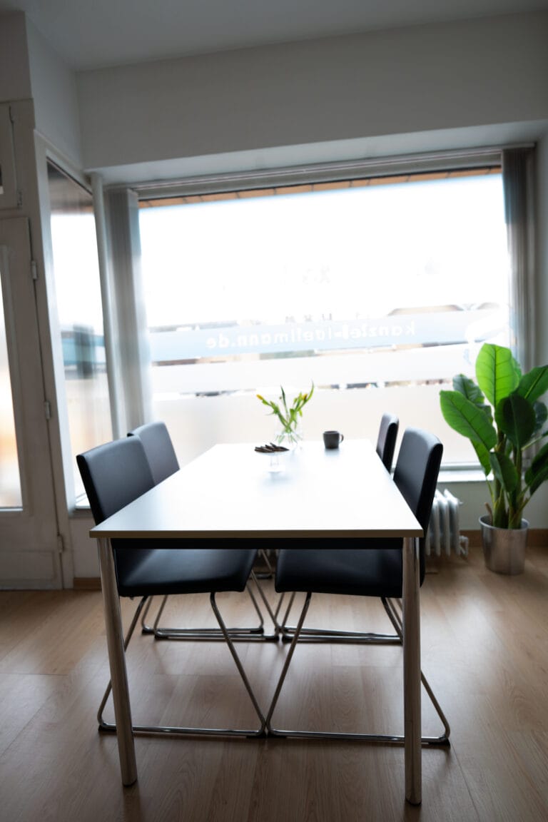 Ein heller Raum mit einem Holztisch und vier schwarzen Stühlen. Der Tisch steht in der Mitte des Raumes und hat eine weiße Tischplatte. Im Hintergrund ist ein großes Fenster zu sehen, das viel Licht hereinlässt.